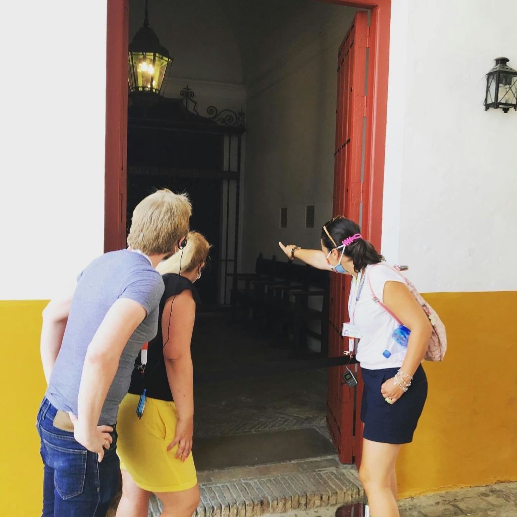 마스크를 착용한 투어 가이드를 포함한 3명이 빨간색 열린 문을 통해 어두운 조명의 방을 들여다보고 있습니다.