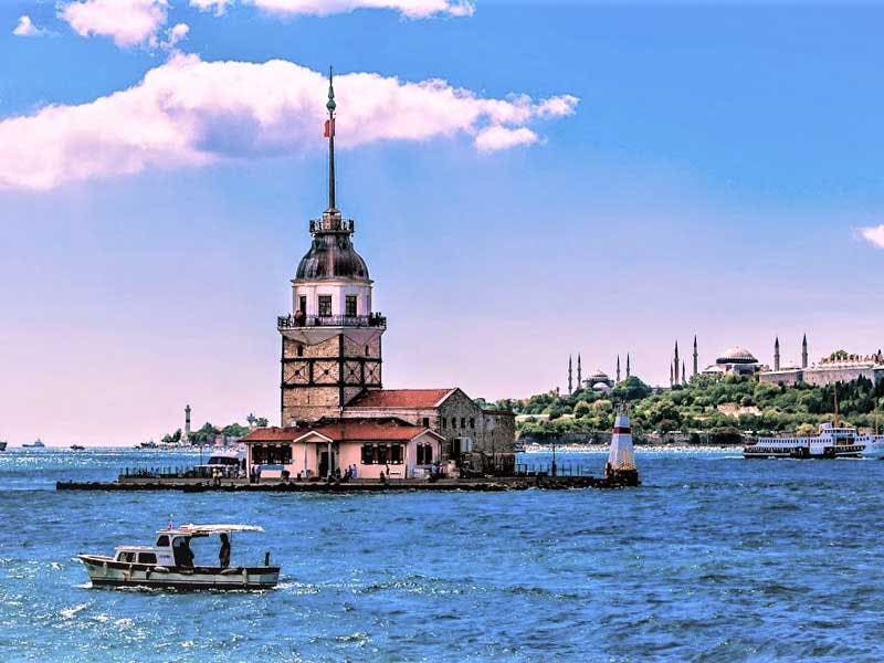 전경에 보트가 있고 푸른 하늘 아래 배경에 도시 풍경이 있는 물줄기의 작은 섬에 있는 역사적인 탑입니다.