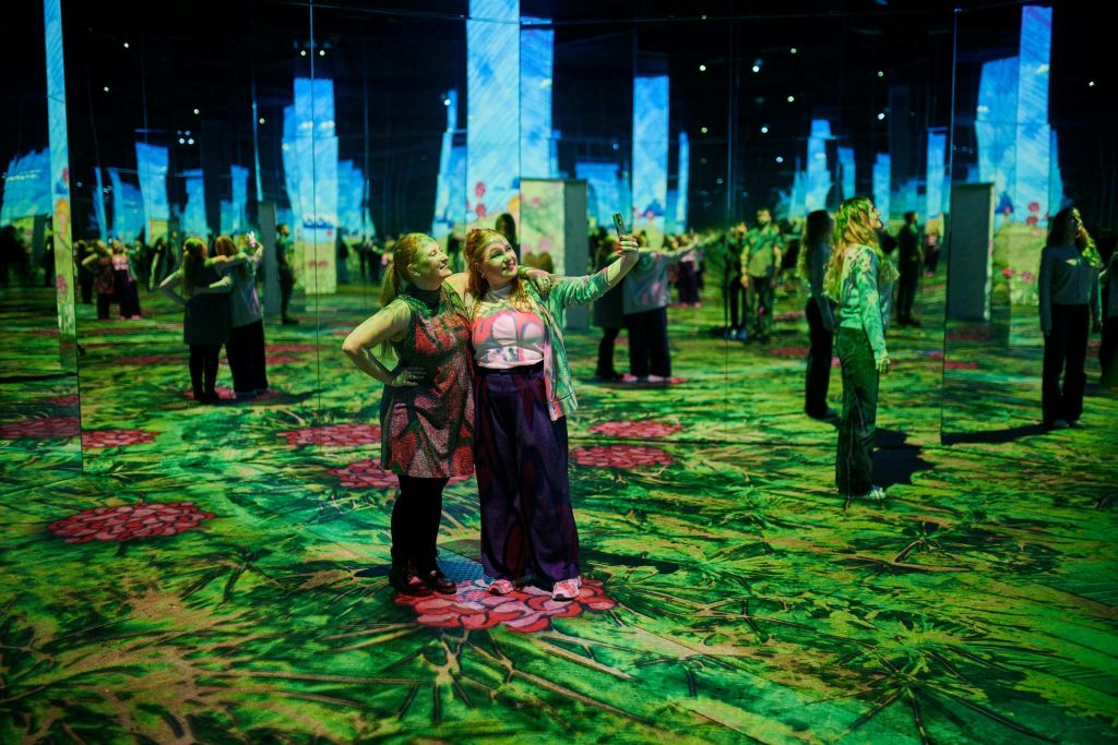 거울 벽과 조명 디자인으로 화려하고 몰입감 넘치는 예술 작품을 설치한 두 여성이 셀카를 찍으며 포즈를 취하고 있습니다.