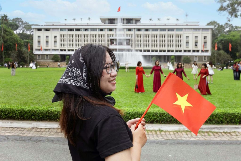 분수가 있는 큰 건물 앞에 베트남 국기를 들고 있는 사람이 서 있습니다. 빨간색 드레스를 입은 사람들이 배경에 있습니다.