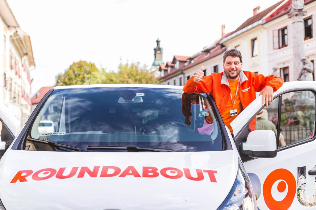 오렌지색 재킷을 입은 남성이 건물과 동상을 배경으로 흰색 밴에 기대어 엄지손가락을 치켜들고 있습니다.