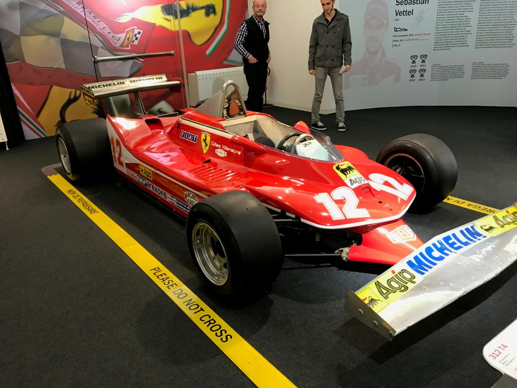 실내에 전시된 후드의 숫자 12가 새겨진 레드 빈티지 포뮬러 1 레이스 카. 두 남자가 그 뒤에 서 있습니다.