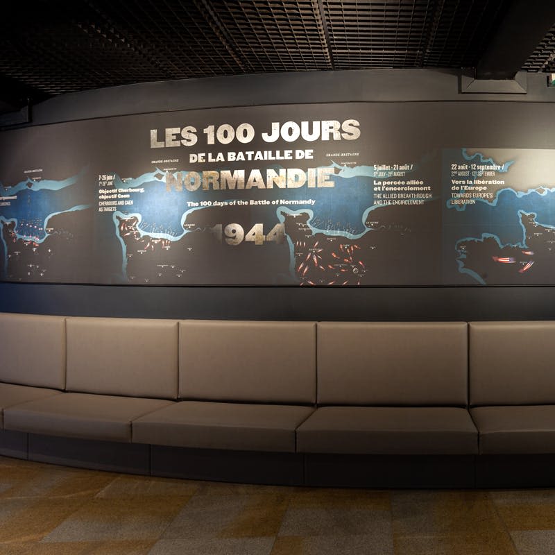 타임라인과 지도와 함께 "Les 100 Jours de la Bataille de Normandie" 를 읽는 보드를 표시합니다. 앞쪽에 곡선 회색 좌석이 있습니다.