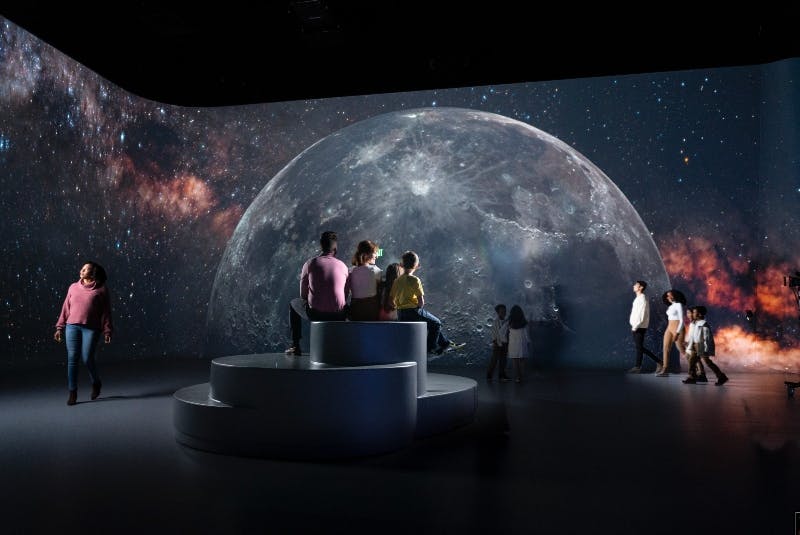 사람들은 어두운 방에서 화면에 별이 있는 크고 사실적인 달 투영을 보며 층이 있는 좌석에 앉아 있습니다.