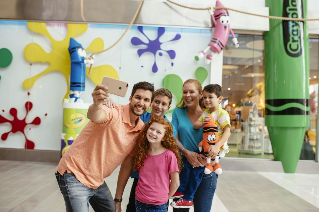 5인 가족이 쇼핑몰에서 화려한 크레용 테마 장식 앞에서 셀카를 찍고 있습니다.