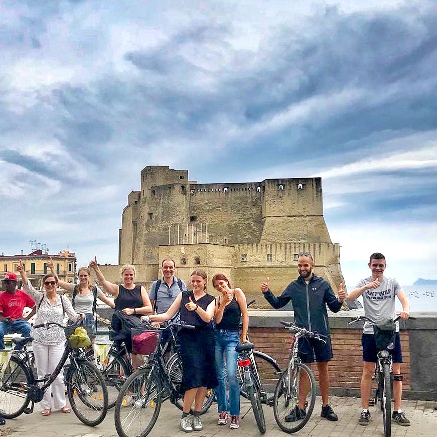 흐린 하늘 아래 역사적인 석조 요새 앞에 자전거를 탄 한 무리의 사람들이 미소를 지으며 서 있습니다.