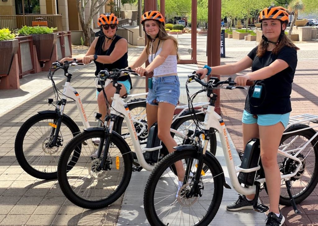 햇살이 내리쬐는 야외 환경에서 포장된 보도에 흰색 자전거가 달린 주황색 헬멧을 쓴 세 사람이 서 있습니다.