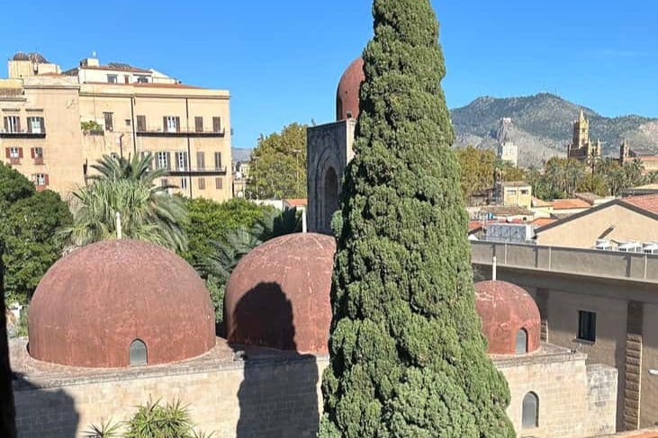 맑고 푸른 하늘 아래 도시 풍경과 산을 배경으로 붉은 돔이 있는 건물 옆에 높고 푸른 편백나무가 서 있습니다.