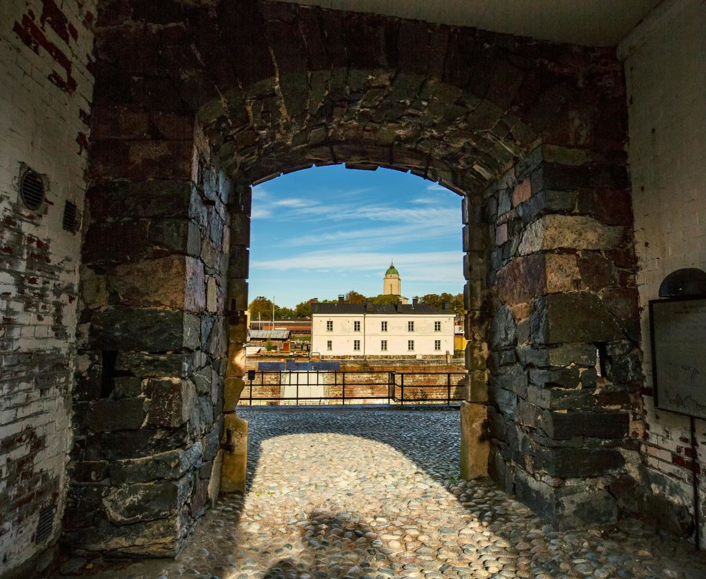 푸른 하늘 아래 녹색 돔이 있는 흰색 건물의 전망을 감상할 수 있는 석조 아치형 통로. 전경의 자갈길.