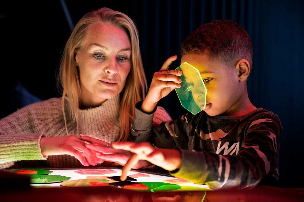 한 여성과 어린이가 연한 테이블 위에 놓인 화려한 반투명 형상과 상호작용하며, 아이는 눈에 초록색 형상을 유지합니다.