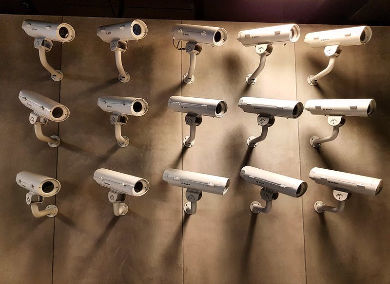 여러 개의 흰색 보안 카메라가 그리드 패턴으로 베이지색 벽에 장착되어 있으며, 모두 다양한 방향을 가리킵니다.