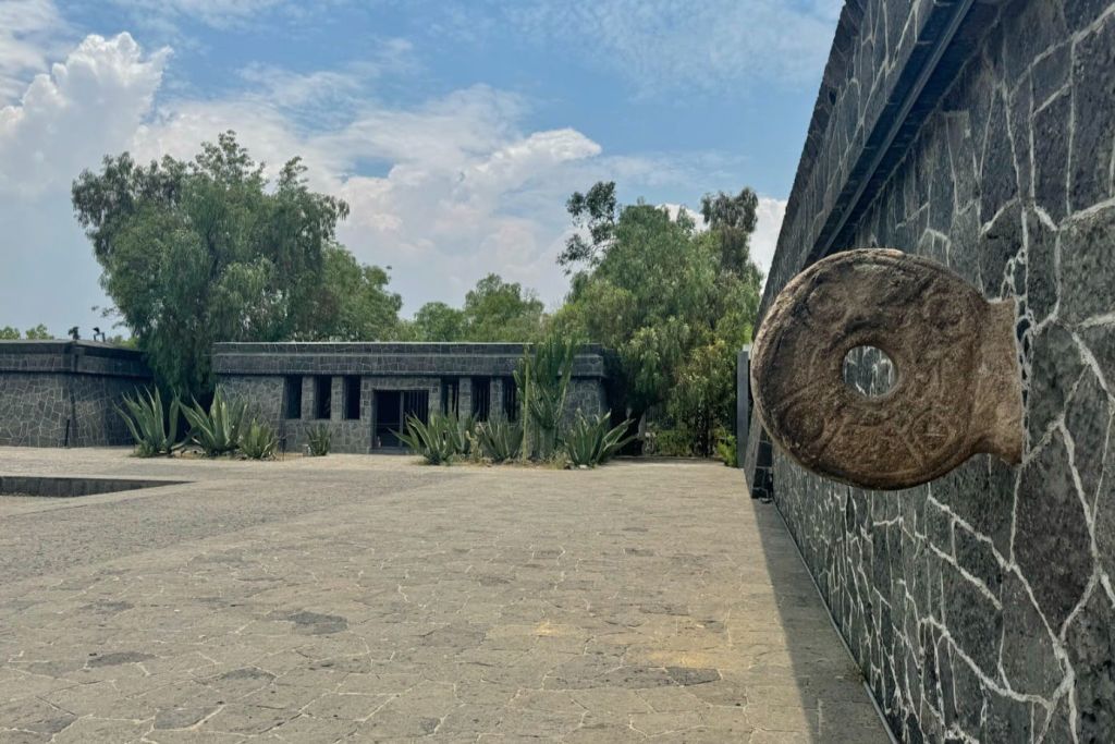 나무로 둘러싸인 벽에 둥근 돌 조각이 있는 돌 마당과 배경에는 작은 돌 건물이 있습니다.