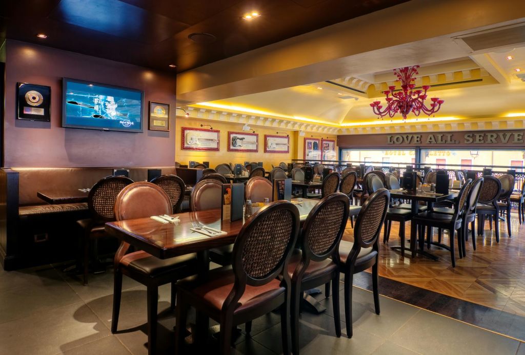 나무 테이블, 갈색 의자, 벽걸이 TV, 액자 사진, 분홍색 샹들리에가 있는 조명이 좋은 레스토랑입니다.