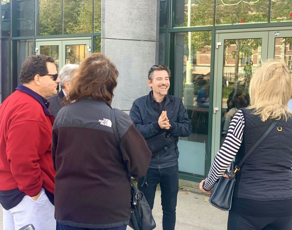 유리창과 문이 있는 건물 밖에서 소수의 사람들과 이야기하면서 웃으며 박수를 치는 남성.