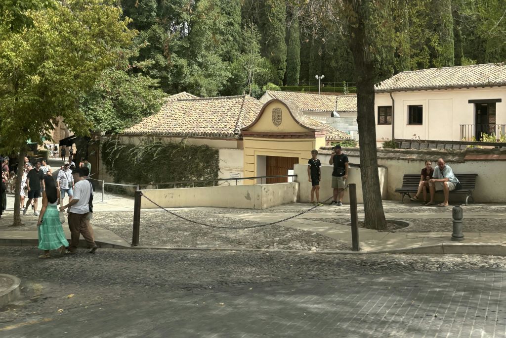 타일로 덮인 지붕과 나무를 배경으로 울타리가 쳐진 건물 입구 근처에 두 사람이 서 있습니다. 산책로가 나란히 휘어집니다.