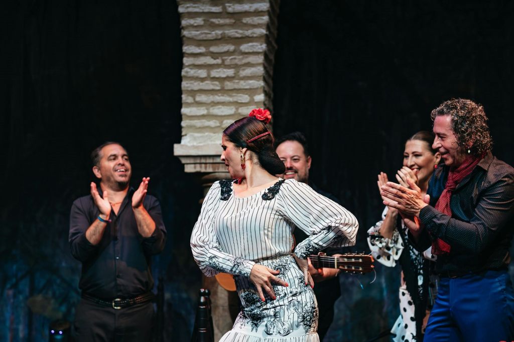 하얀 드레스를 입은 플라멩코 댄서가 무대에서 뮤지션을 배경으로 박수를 치며 공연을 펼칩니다.