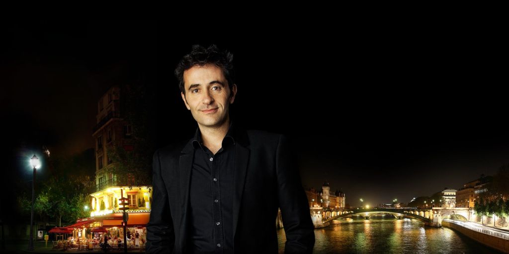 검은 양복을 입은 남성이 어두운 배경에 서서 왼쪽에는 불이 켜진 거리 풍경이, 오른쪽에는 강이 있습니다.