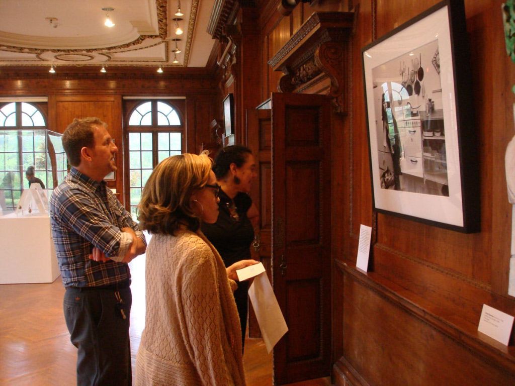 나무 패널로 꾸며진 갤러리에서 아치형 창문을 배경으로 액자 모양의 예술 작품을 감상하는 세 사람.