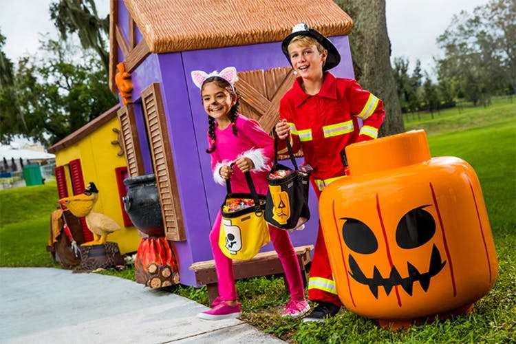 할로윈 의상을 입은 두 명의 어린이, 분홍색 고양이와 소방관이 장난스럽고 으스스한 집 앞에서 사탕 양동이를 들고 있습니다.