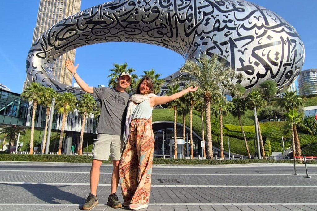 아랍 서예로 장식된 미래형 금속 조각상 앞에서 두 팔을 들어 올린 채 포즈를 취하는 남성과 여성.