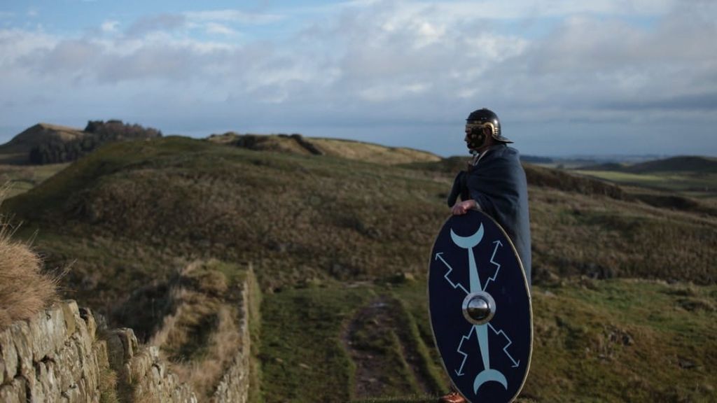 헬멧과 망토를 착용한 사람이 흐린 하늘 아래 잔디 언덕 꼭대기에 서 있는 동안 방패를 들고 있습니다.