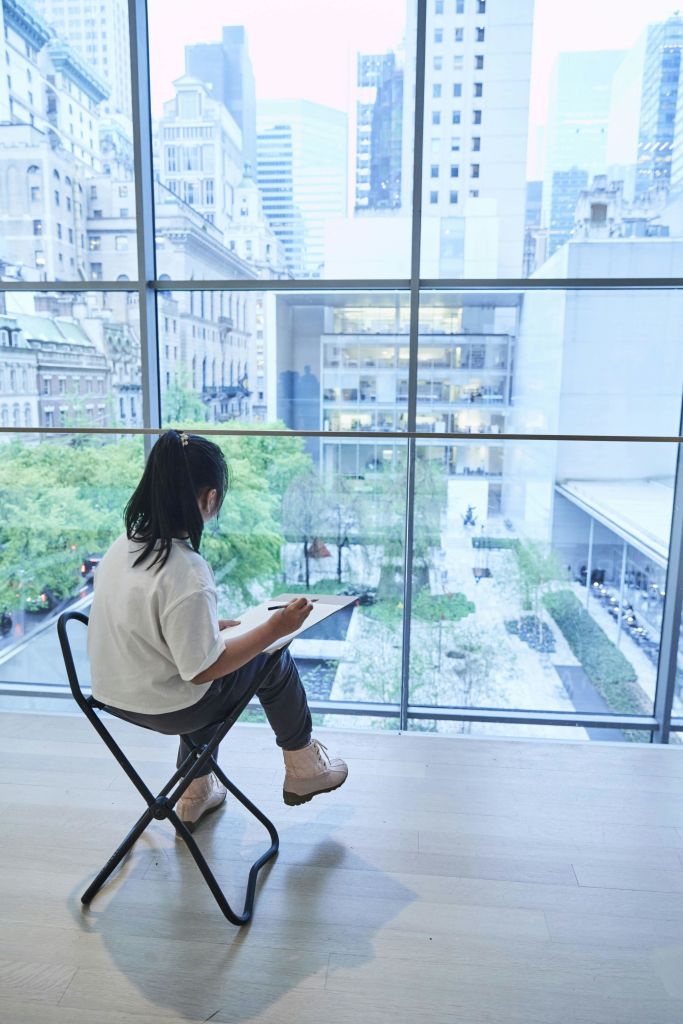 한 사람이 의자에 앉아 커다란 패드에 스케치를 하고 나무와 건물이 있는 도심 안뜰이 내려다보이는 벽 크기의 창문을 마주하고 있습니다.