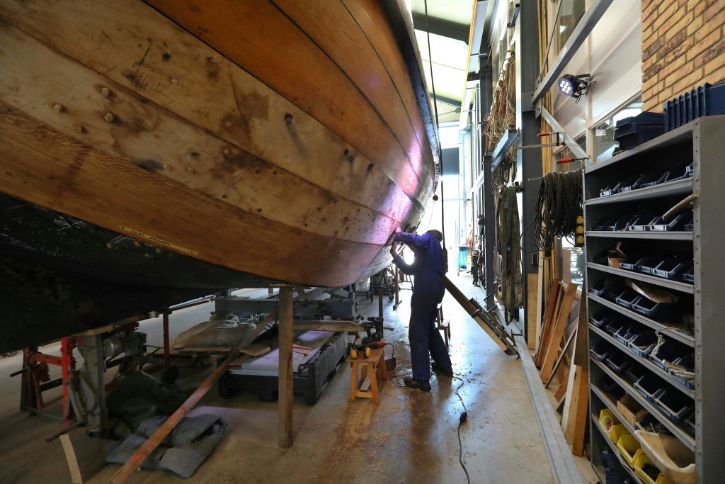 작업장에서 도구와 장비로 둘러싸인 보트의 나무 선체에서 작업하는 사람.