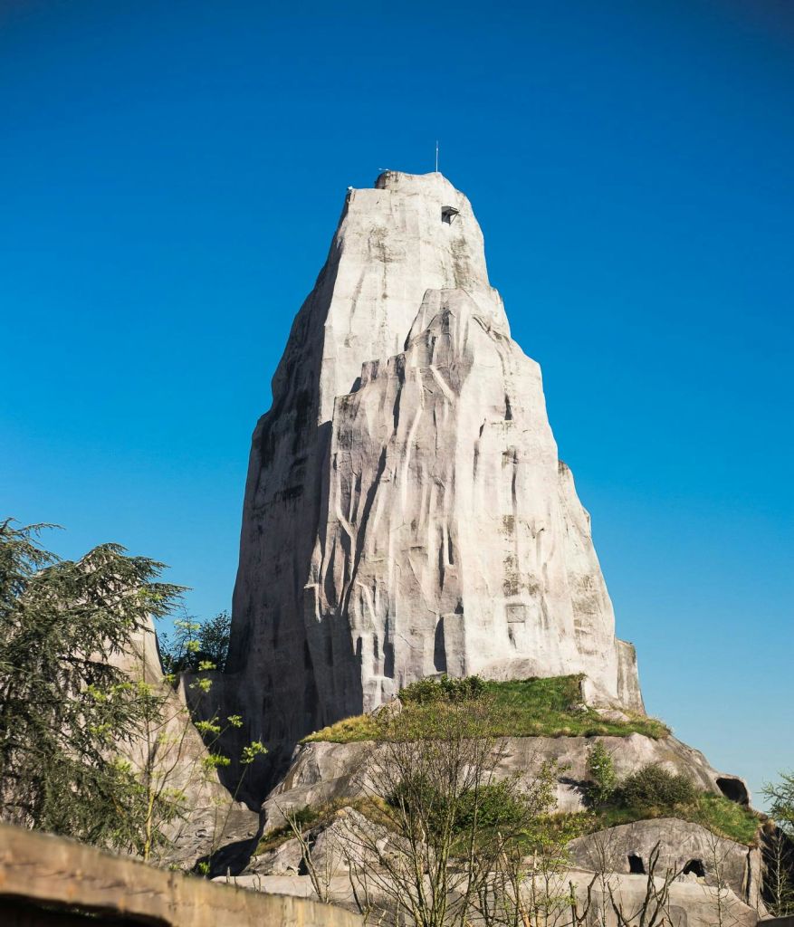 청명한 푸른 하늘 아래 희박한 녹지와 관목으로 둘러싸인 높고 바위투성이의 산악 구조물.