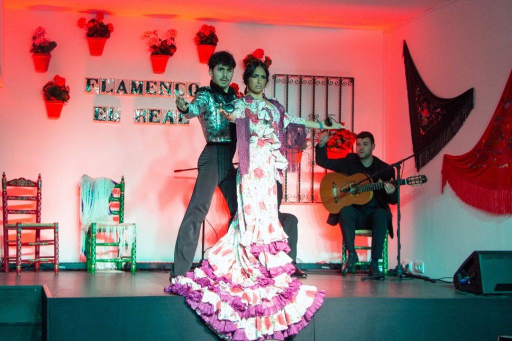 전통 복장을 한 두 명의 플라멩코 무용수가 무대에서 공연하고, 배경에 앉아 있는 기타리스트가 함께합니다.