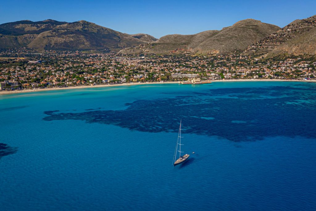 맑은 하늘 아래 건물과 산을 배경으로 해안선 근처의 맑고 푸른 바다에서 요트를 타보세요.