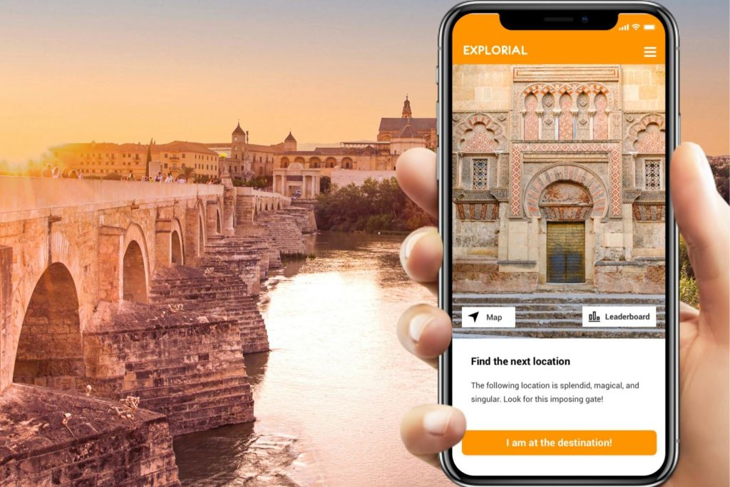 Explorial 앱이 표시된 스마트폰을 들고 있는 손은 역사적인 장소를 보여줍니다. 석양을 배경으로 한 돌다리가 강에 걸쳐 있습니다.