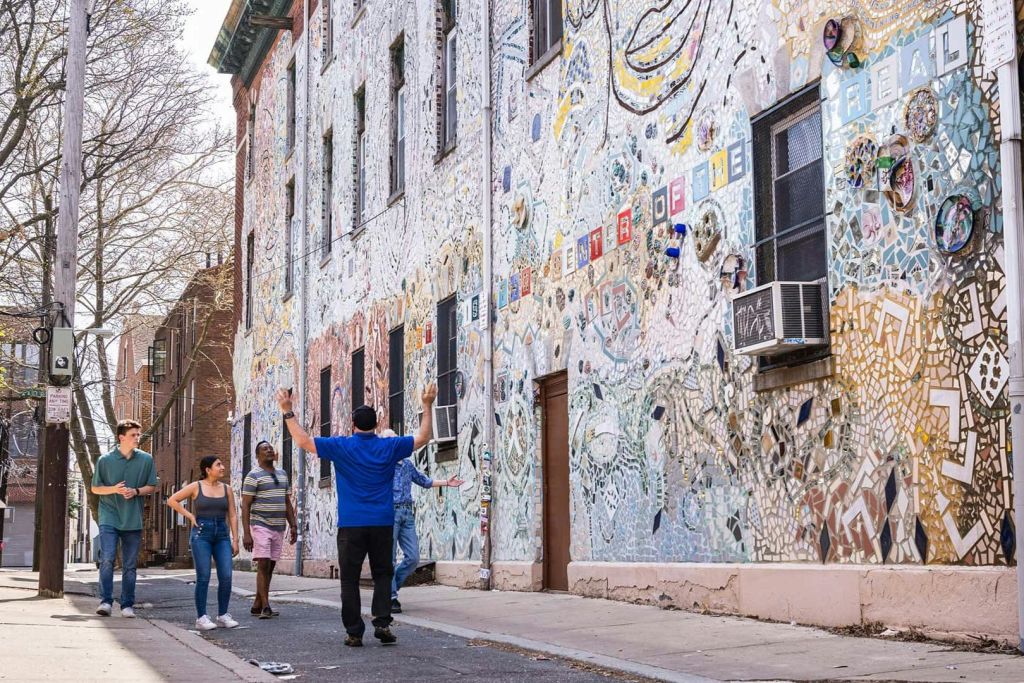 창문과 에어컨이 있는 건물의 화려한 모자이크가 덮인 벽 옆 거리를 걷는 사람들.