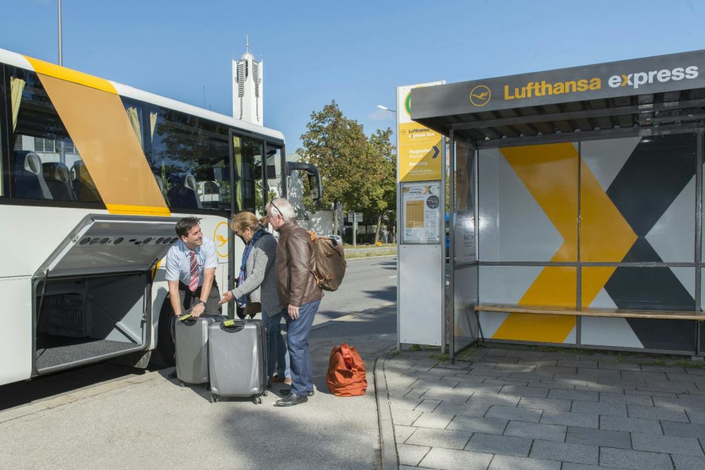 여행 가방을 든 두 명의 여행자가 주차된 버스 근처에서 버스 기사와 이야기를 나누고 있습니다. 빨간색 배낭이 바닥에 놓여 있습니다.