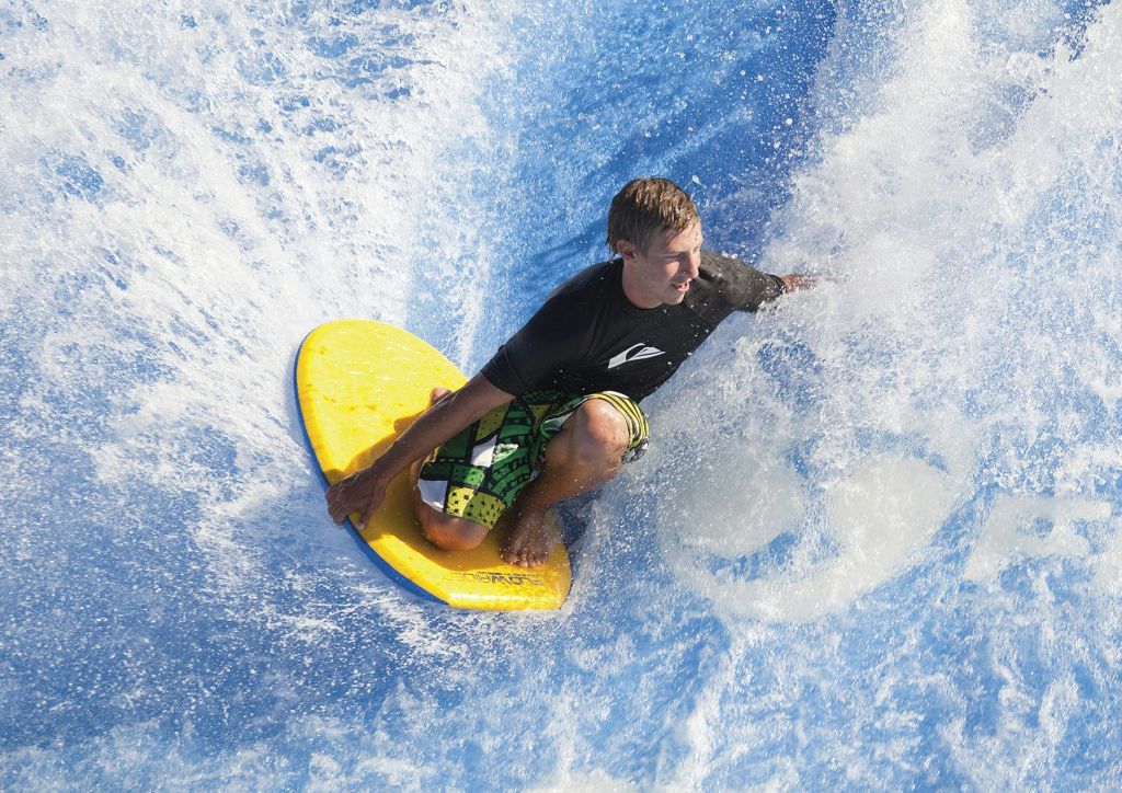 노란색 보디보드에 무릎을 꿇은 사람이 워터파크에서 검은색 발진 가드와 녹색 패턴의 반바지를 입고 파도를 타고 있습니다.
