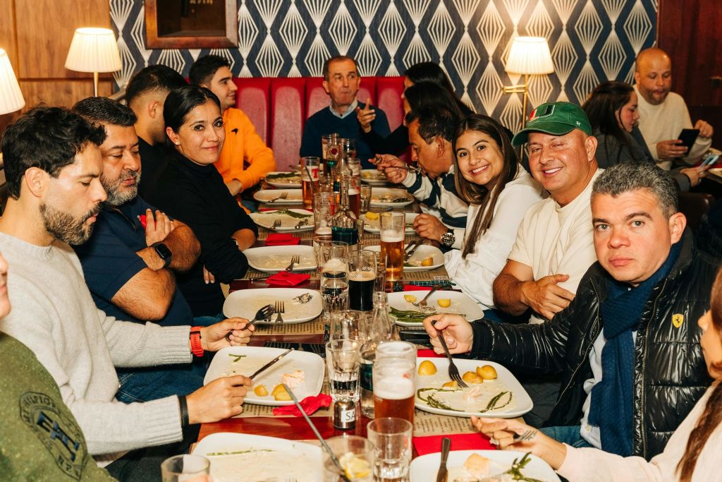 한 무리의 사람들이 긴 식탁에 둘러앉아 음식과 음료, 전등을 배경으로 앉아 있습니다.