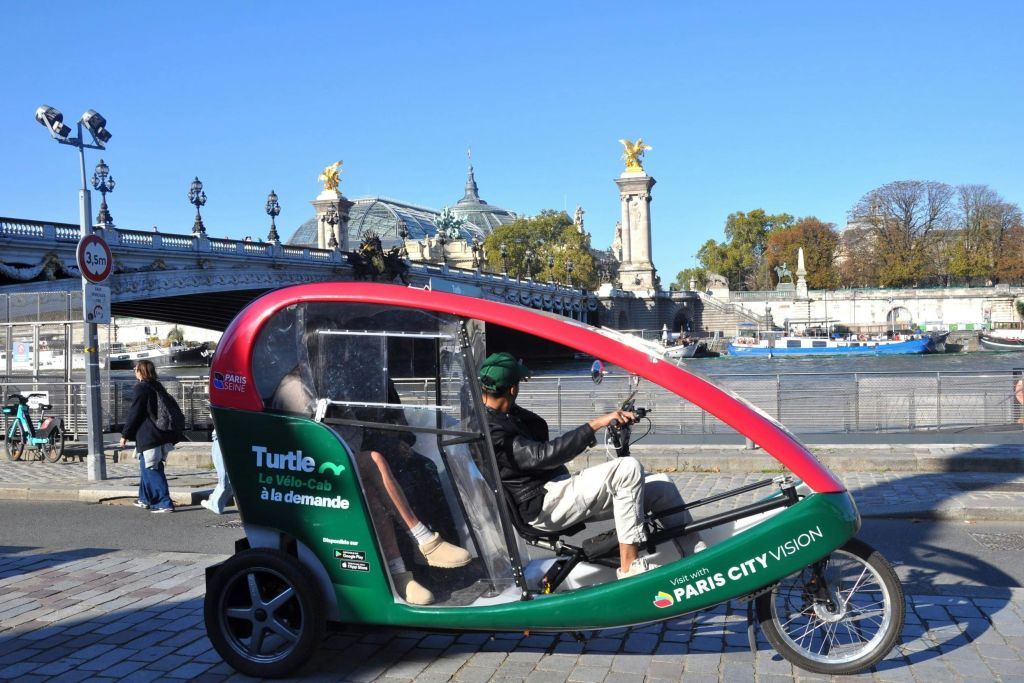 도시 분위기의 조각상으로 장식된 다리 근처에서 '파리 시티 비전 (Paris City Vision)' 브랜드가 새겨진 초록색과 빨간색 페디캡을 타는 사람이 있습니다.
