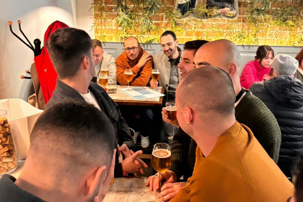 벽에 식물이 걸려 있는 따뜻하게 빛나는 펍에서 각자 맥주 한 잔을 들고 무심코 수다를 떨며 웃고 있는 한 무리의 남자들.