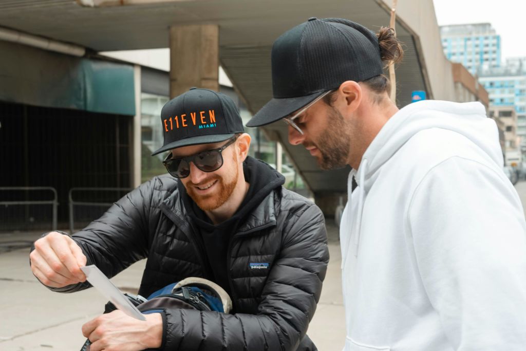 선글라스와 모자를 쓴 두 명의 남성, 한 명은 검은색 재킷을 입고 다른 한 명은 흰색 후드티를 입은 채 도심 속에서 종이를 바라보고 있습니다.