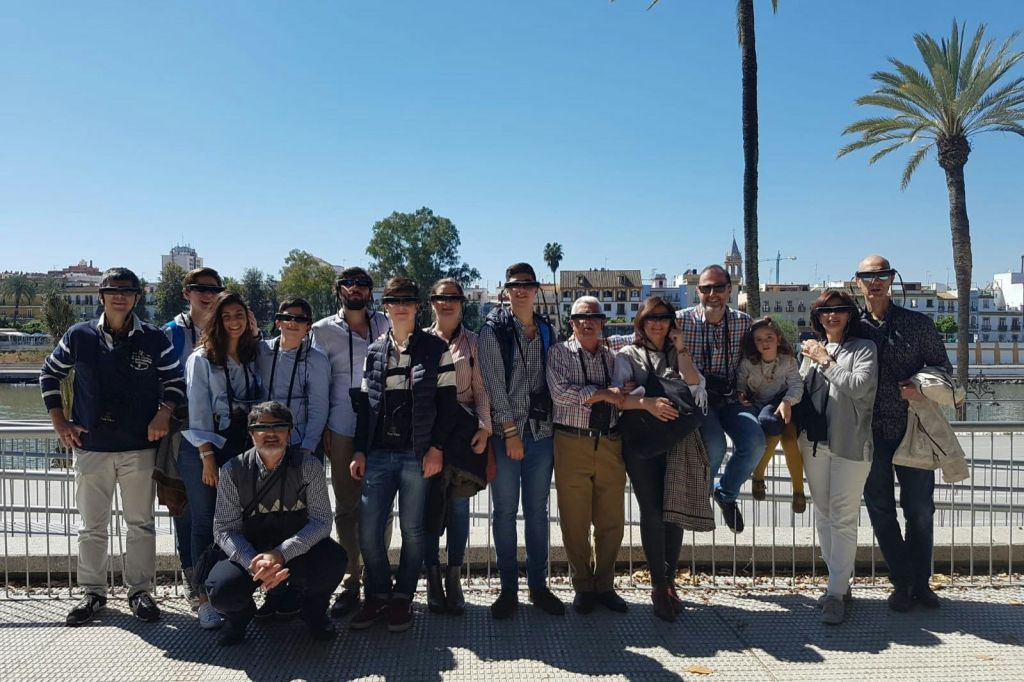 맑고 푸른 하늘 아래 야자수와 건물이 있는 해안가 도시 풍경 앞에서 포즈를 취하며 함께 서 있는 사람들의 그룹.