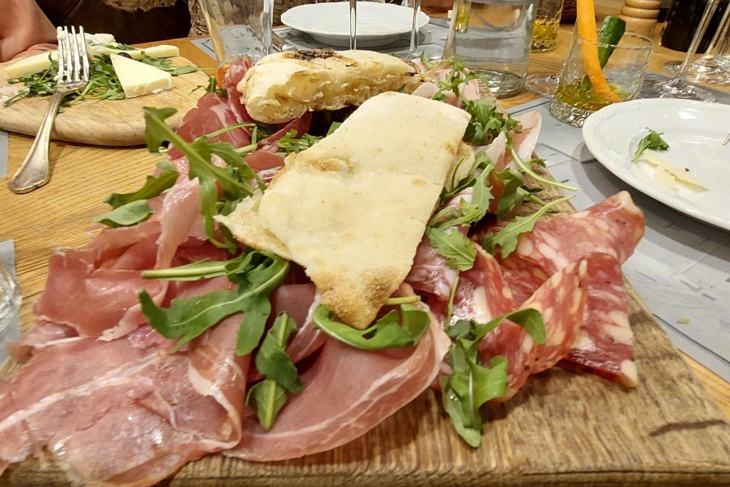 테이블 위에 접시와 유리잔으로 둘러싸인 다양한 경화 고기, 신선한 아루굴라, 평평한 빵이 있는 나무 판입니다.