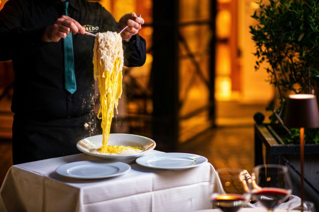 어두운 제복을 입은 사람이 하얀 접시와 하얀 식탁보가 놓인 테이블에서 치즈로 덮인 파스타를 제공합니다.
