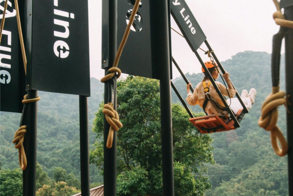 '스카이 라인' 이라고 적힌 커다란 그네 세트에 헬멧을 쓴 사람이 울창하고 푸른 산을 배경으로 그네를 타고 있습니다.