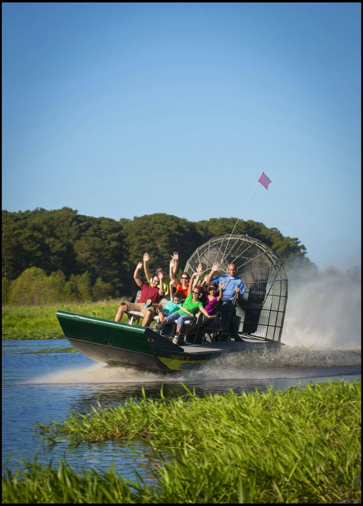 한 무리의 사람들이 에어보트를 타고 잔디가 우거진 수로를 가로질러 나무를 배경으로 흥분한 듯 손을 들고 있습니다.