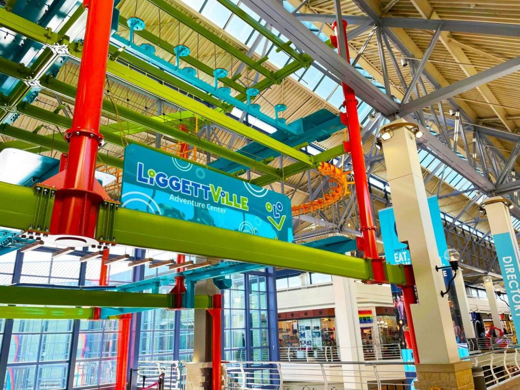 녹색, 빨간색, 파란색 구조의 다채로운 로프 코스가 있는 실내 어드벤처 센터와 "LiGGEtTViLLE Adventure Center" 표지판이 있습니다.