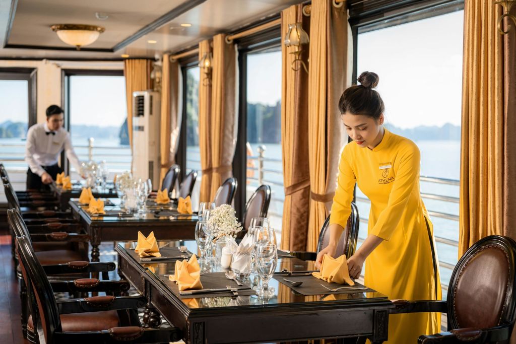 노란색 유니폼을 입은 여성이 아름다운 수변 전망을 감상할 수 있는 커다란 창문 옆 식탁에 테이블을 배치하고 있습니다.