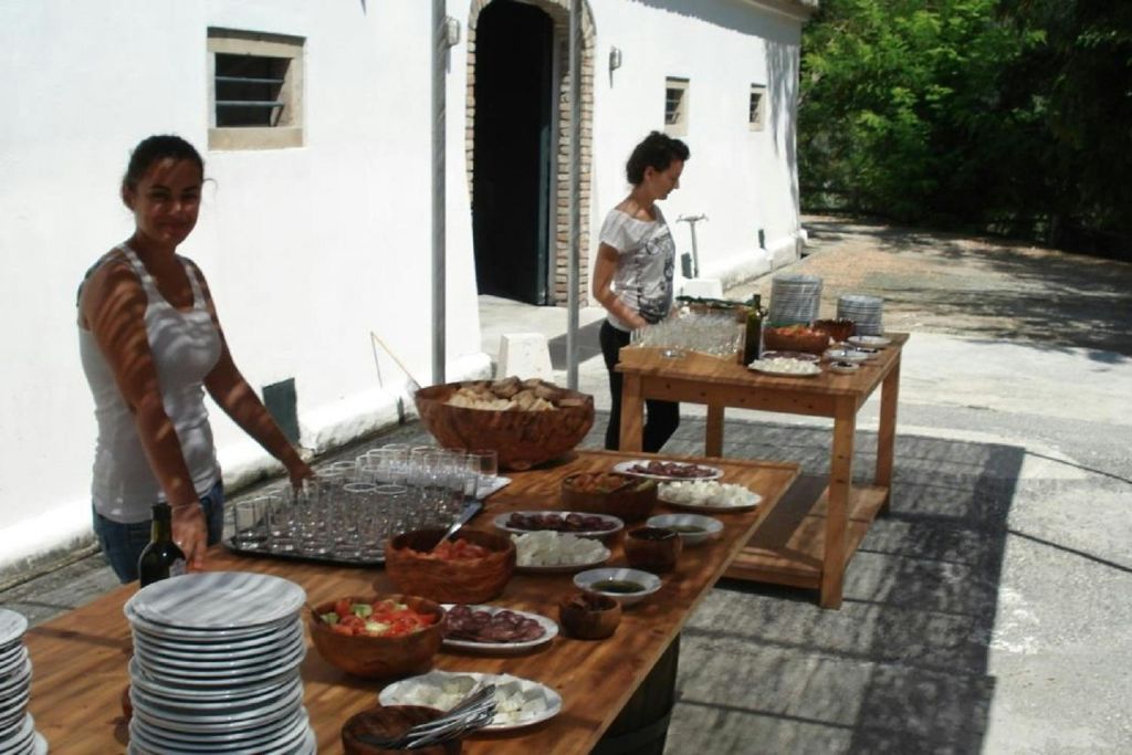 흰색 건물 옆 야외에는 접시, 유리잔, 음식 그릇이 가득한 테이블 옆에 두 명의 여성이 서 있습니다.