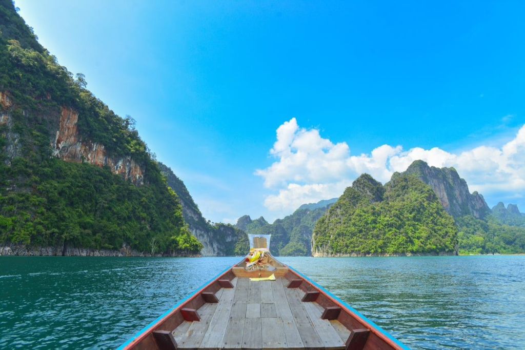 구름이 흩어져 있는 맑은 하늘 아래 푸른 호수의 푸르고 바위가 많은 언덕 사이로 롱테일 보트가 이동합니다.
