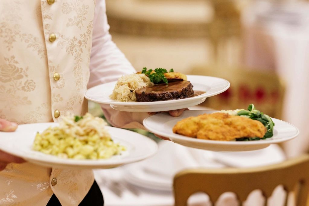 흰색과 금색 조끼를 입은 웨이터가 파스타, 스테이크, 튀김 요리를 포함한 고급 음식을 제공합니다.