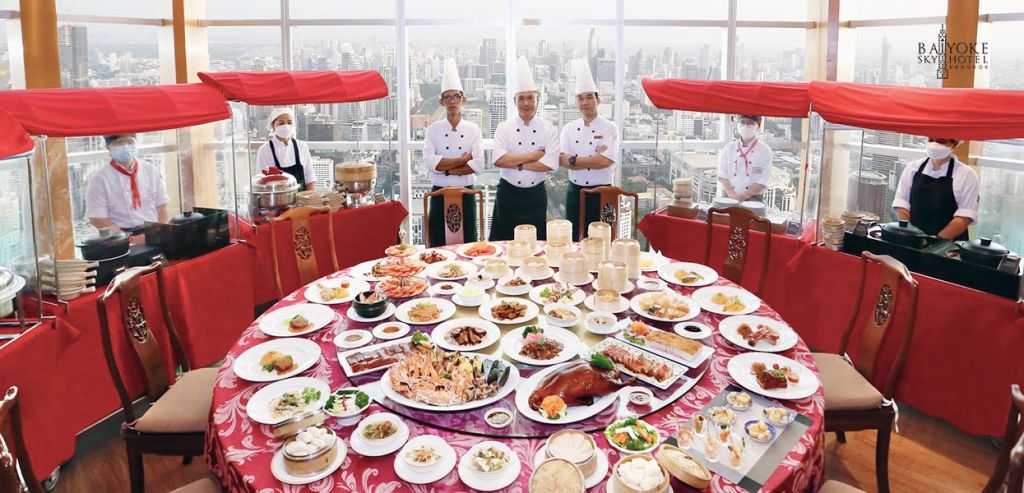 네 명의 셰프가 다양한 요리가 가득한 커다란 원형 테이블 뒤에 서서 커다란 창문을 통해 도시 풍경을 내려다보고 있습니다.