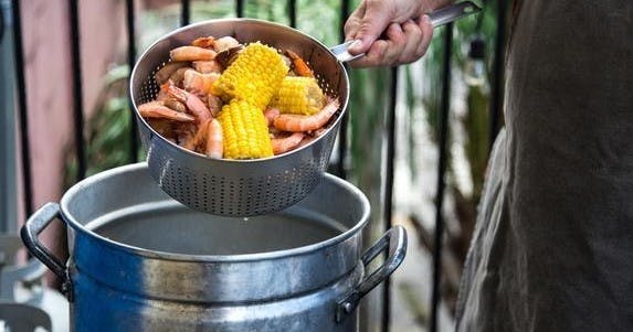 옥외에서 끓는 냄비 위에 새우와 옥수수를 올린 채로 콜랜더를 들고 있는 손.